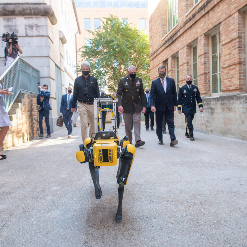 Boston Dynamics Robot walks down the sidewalk with group of people including Army men and researchers behind